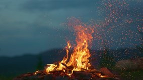 Campfire at dusk against the backdrop of mountains and forest. Tourist bonfire with sparks flying in the air, slow motion shot. - Powered by Shutterstock - Get 15% off with code: PIKWIZARD15