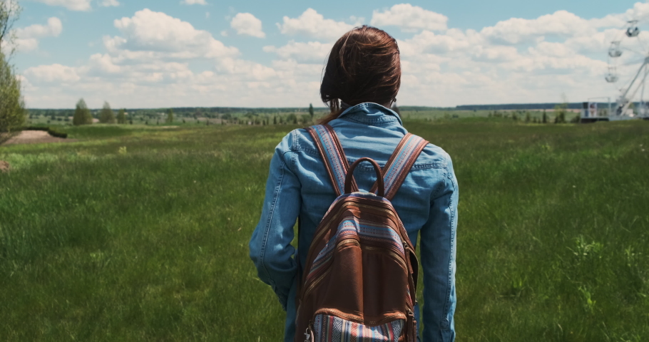 Young woman with backpack dressed in denim walks through green field with tall grass. Rear view, camera follows girl. White clouds blue sky, nature, summertime, daytime.