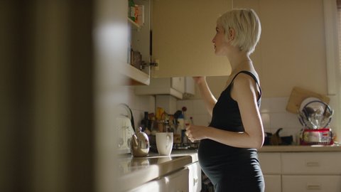 Young pregnant female in her kitchen opening her cupboards to start making a hot drink, in slow motion