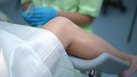 Close-up leg of woman lying on gynecological chair with blurred doctor using vaginal speculum at background. Expert obstetrician examining Caucasian young patient in hospital