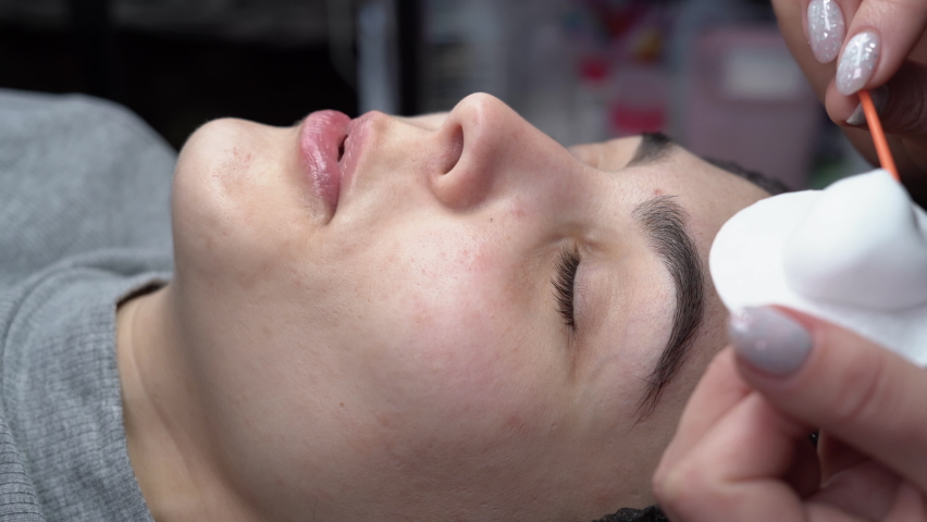 A young woman undergoes an eyelash extension procedure and removes mascara with a cotton swab and stick in a beauty salon. Eyelid preparation. | Shutterstock HD Video #1091298205