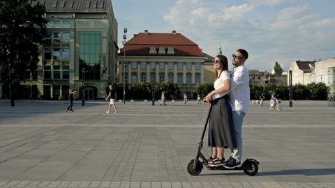 Young man rides his girlfriend on an electric scooter around the city square. Happy girl laughs and spreads her arms to the sides.