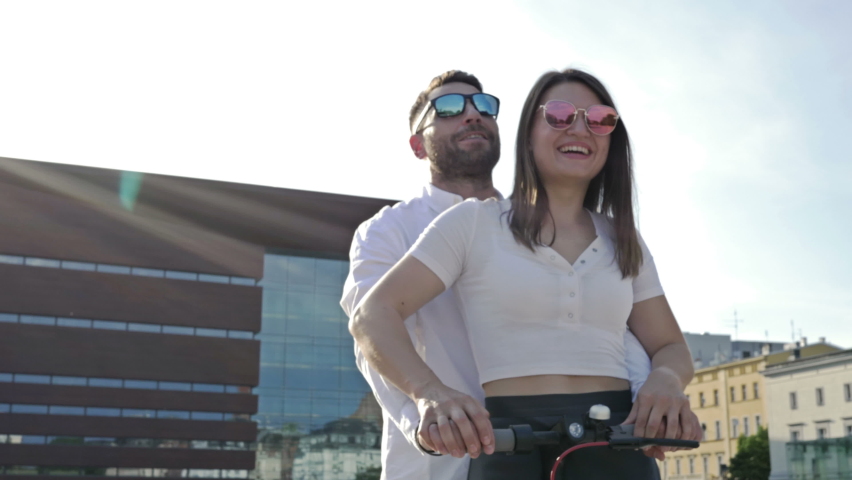 Couple of lovers ride an electric scooter through a beautiful square of a European city. Man and woman in sunglasses smile happily.