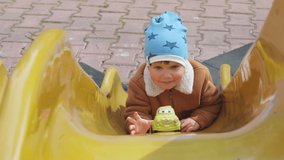 Cheerful funny little boy in blue hat and warm clothes plays on playground in kindergarten and rolls his yellow toy car on bright multi-colored slide, in cloudy autumn weather. HD slow-motion video