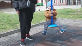 Little cheerful boy in blue hat and warm clothes spends his free time on bright playground in kindergarten in autumn empty park, and rides on blue modern swing in cloudy weather. HD slow-motion video