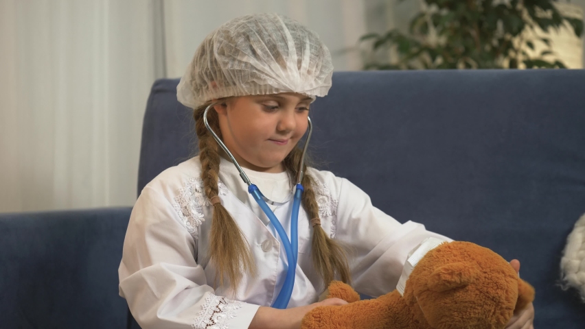 Little girl in white doctor's coat is playing. child plays doctor. girl using stethoscope treating teddy bear sitting in an isolated room. small child dreams of becoming doctor by helping teddy bear