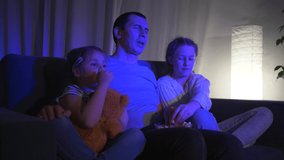 Happy family is sitting on the couch in the evening, watching TV, a projector. Children watch an interesting movie with their dad. Cheerful family eating popcorn in the evening in front of the TV.