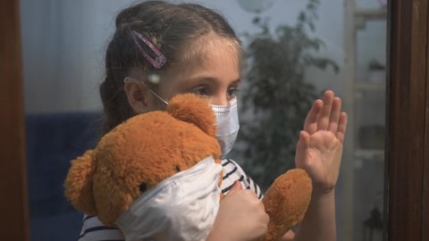 Protection of children, medical. Sad girl in protective medical mask at window. girl is alone at home with toy. sad child in protective mask in hospital looks out window. child is isolated at home.