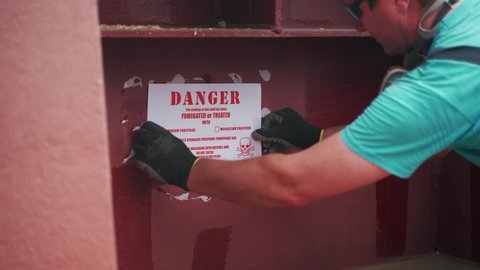 2021-06-09 Mariupol, Ukraine. Ukrtransagro LLC. Shipping pest control worker sticking biohazard danger sign on shipping container or cargo holds of bulker grain ship in seaport after fumigation.