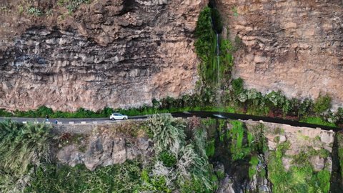 White car drives under Angels waterfall that cascades onto cliffside road; drone