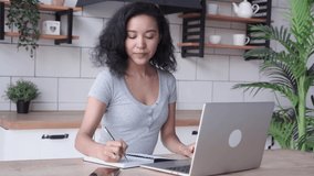 African American woman sits indoor and using app on laptop for video call to friends, making sketches in notebook. Young female employee in casual wear talking online via video connection waving hands
