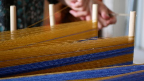 Winding a warp for the weaving loom using warping frame and blue and yellow yarns. Wooden pegs and colored threads, selective focus