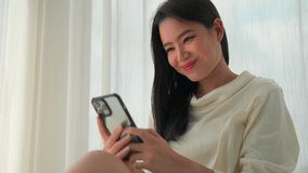 Happy young woman using smartphone to play social media in the room talking with friends with beautiful smile and happy face relaxing at home happily enjoying with modesty weekend time