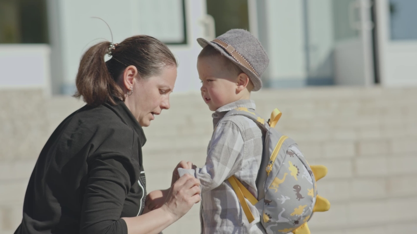 back to school concept, mother sending baby boy child to school first time. caucasian female checking clothes and school bag of preschooler kid, hugs her kid, waving hands, encouraging son in front of