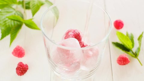 Raspberry summer cocktail. Summer drink.Splashes and drops of a refreshing cocktail Mineral water is poured into a glass with ice cubes and raspberries on a light wooden table. 4k footage