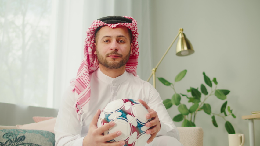 Middle eastern football fan supporting favorite team, watching match on TV. Young man holding soccer ball. Wearing traditional Islamic male clothes. Worship and culture concept.
