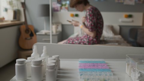 Selective focus of white pill jars and pill organizer on table by bed in hospital room at daytime, blurred girl sitting on background
