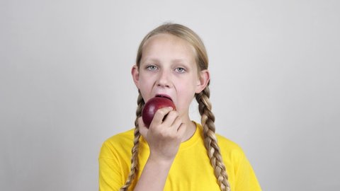 portrait of a cute little girl with braided pigtails biting and eating a ripe red apple, looking at the camera in a yellow t-shirt on a white background. Healthy food and beauty. diet
