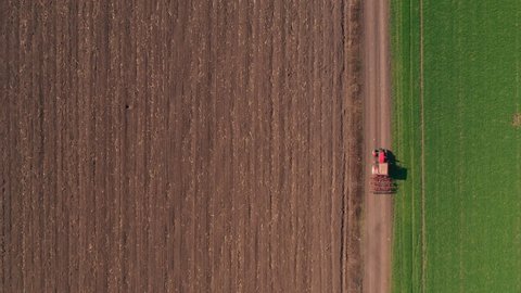Aerial view of agricultural tractor with tillage equipment attached on dirt road through cultivated landscape, drone pov directly above