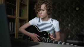 Adorable curly little girl learning to play the guitar in virtual meeting for play music online together with friend or teacher in video conference with laptop. Shooting in slow motion.