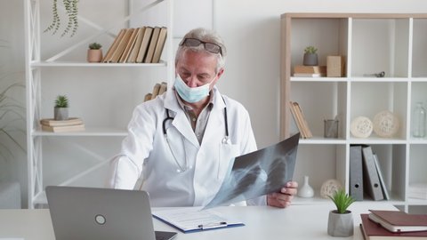 Lung radiology. Respiratory infection. Coronavirus infection. Middle-aged male doctor in face mask studying chest xray scan at modern clinic workplace interior.