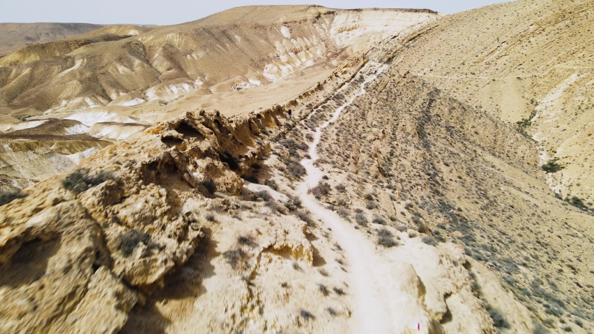 Aerial flying low over ridge and hiker on desert trail showing spectacular landscape on sunny day with blue sky. Location is Ramon Crater in the Negev Desert, Israel.