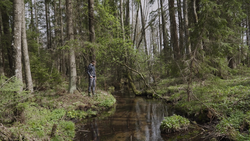 Lonely slender man leaning on wooden twig, approaches edge of river bank in wild forest, carefully descends into water in rubber boots. Bed of spring water on sunny day. Guy is relaxing, slow motion.