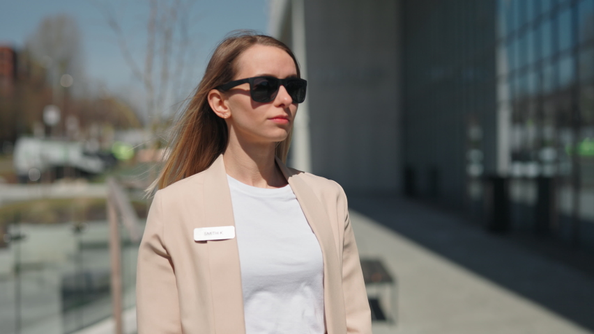 Charming young manager smiling and looking at camera while standing near office center with crossed arms. Beautiful business woman wearing stylish formal jacket and trendy sunglasses.