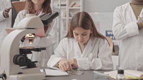 Medium slowmo of multiethnic pupils in lab coats examining unrecognizable sample on petri dish looking at it under microscope during Science class - Powered by Shutterstock - Get 15% off with code: PIKWIZARD15