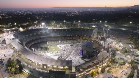 People gathering to sport game or music concert. Aerial illuminated Dodgers Stadium with night LA cityscape on background. Close up drone panorama around Dodgers Stadium, Los Angeles USA, June 2022