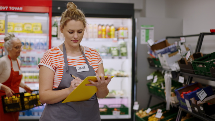 Young shop assistant in supermarket in vegetable shell, in bakcground is her colleague filling stock.