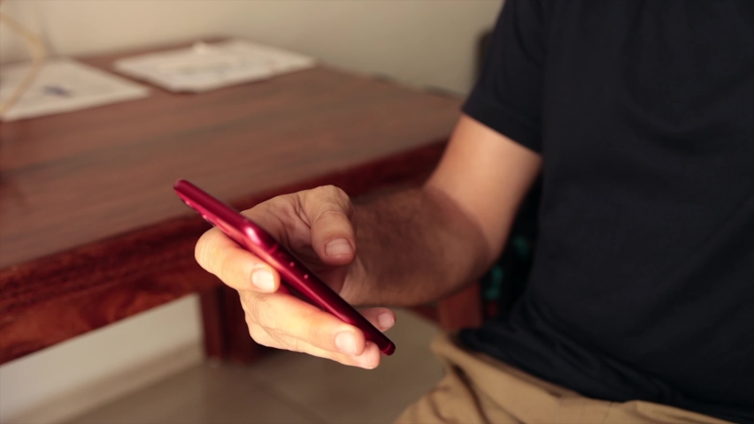White caucasian hairy man scrolling and typing on his cellular phone with wood furniture behind with bright sunlight