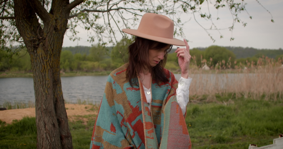 Woman in a hat and poncho at cloudy autumn day stands on countryside with river.