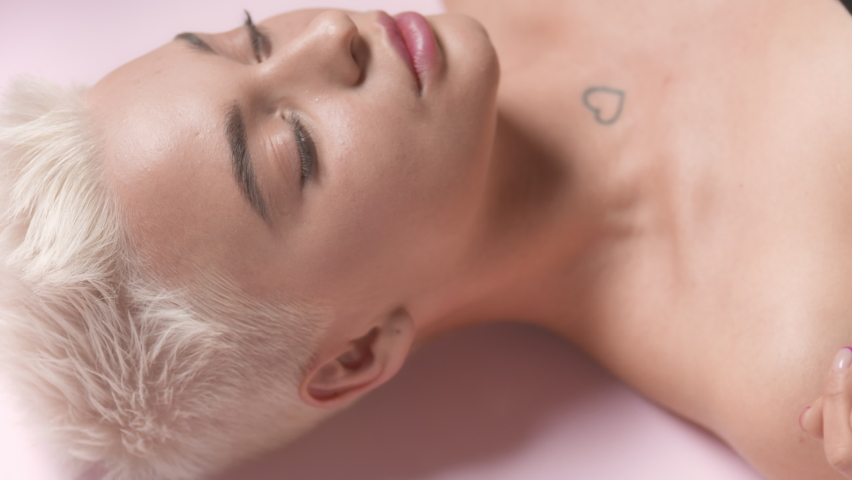 Good-looking young slim Caucasian woman with short light hair turns her head to the camera, opens her eyes and strokes her face through rippling water on pale pink background | Moisturizing commercial