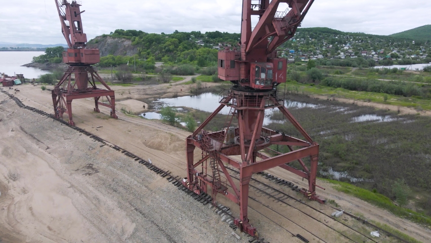 Aerial view of an old abandoned burgundy cargo ship close to crane stands at the bank of a river in a hollow near a village on the hills on a sunny summer evening