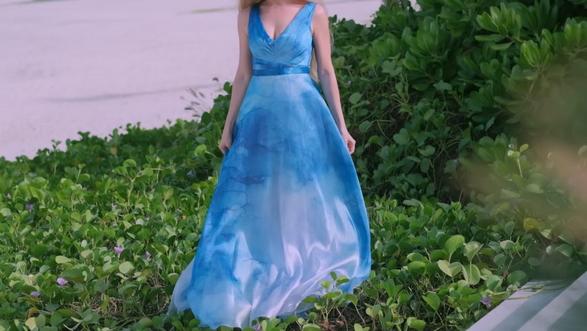 Blonde girl wearing long blue summer dress stays on the grass fragment on the shore, slow motion