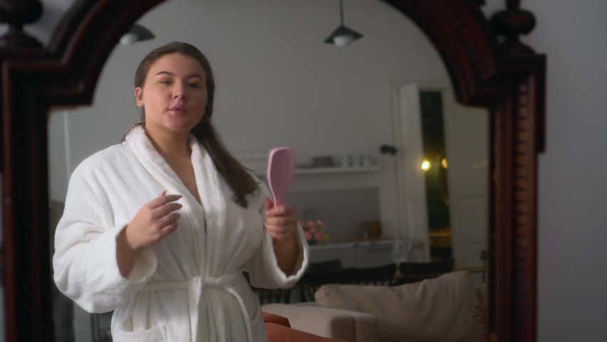 Young overweight woman is singing with comb in hand and dancing in home interior spbd. Close view of beautiful Caucasian female looks in mirror and sings with smile, moves her body actively and stands