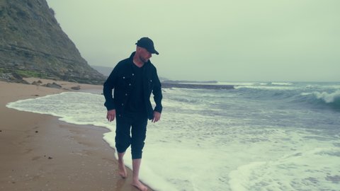 Barefoot  man walks on the empty ocean beach on the summer cloudy rainy weather. Sad adult man walks along a sandy beach at high tide. Human feels loneliness and melancholy, outdoors. 4k cinematic