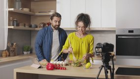 Emotional caucasian couple gesturing and talking while recording video on camera during cooking process on kitchen. Young bloggers sharing with their subscribers recipe of healthy meal.