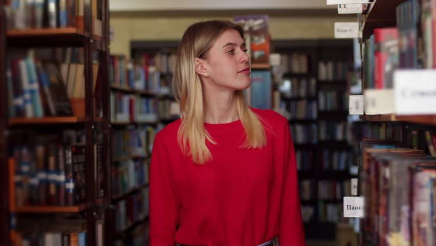 Caucasian blonde woman in a red sweater is standing in the library among the shelves with books, looking around and looking into camera, portrait