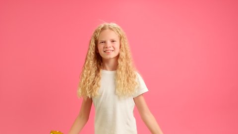 Happy smiling 16 year old teenage girl in white T-shirt is having fun at party, covers her face with golden inscription party, invites everyone to party or birthday standing on pink studio background.