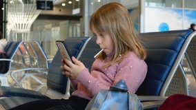 Cute little kid girl looks video on smartphone in airport. Internet concept or wireless technology concept