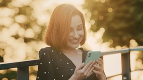 Attractive redhead girl teenager woman stands on street outdoors sunlight rays sunset looks in smartphone screen use mobile smiling watch funny video in internet chatting with boyfriend read good news