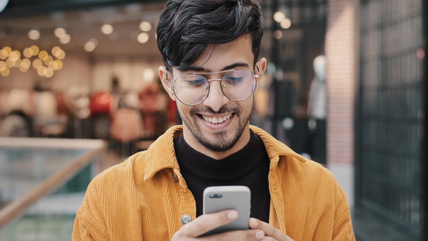Close-up happy indian guy laughs watching funny video on smartphone looking at phone screen browsing web page on social network young man typing sms on dating site remote chat using virtual service | Shutterstock HD Video #1091374511