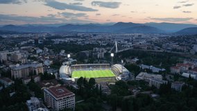 Cityscape featuring a stadium with green grass soccer field (football pitch) illuminated by floodlights at night, in the dark city of Podgorica, Montenegro, under colorful sunset sky in the evening. - Powered by Shutterstock - Get 15% off with code: PIKWIZARD15