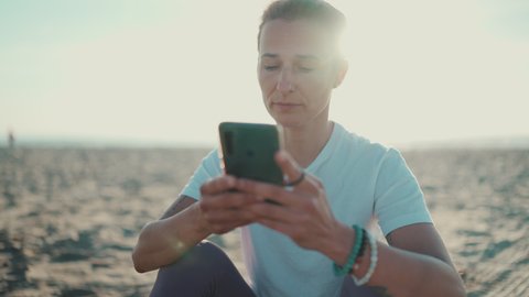 Close up shot of beautiful woman checking her smartphone resting after workout on the beach. Modern technology