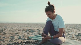Beautiful woman sitting on yoga mat checking her smartphone on the beach. Young yogi woman resting with mobile phone by the sea