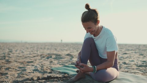 Beautiful woman sitting on yoga mat checking her smartphone on the beach. Young yogi woman resting with mobile phone by the sea