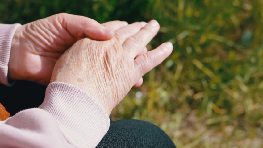 Elderly Woman Rubbing Wrinkled Hands, Sitting in Nature in the Rays of Sunlight. A worried retired woman is nervous, warming hands in sun in the park. Pensioner thinking about social issues outdoors. Royalty-Free Stock Footage #1091384319