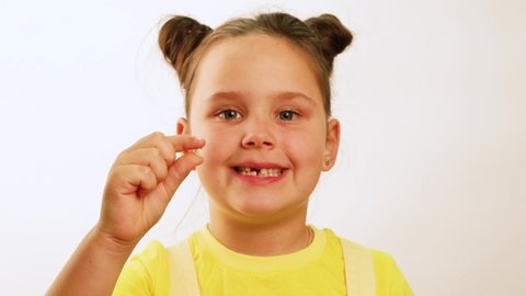 Little funny girl smile and emotionally point finger at gap in mouth from fallen tooth closeup, white background. Hold and show white milk tooth in hand. Concept of healthy teeth and beautiful smile.